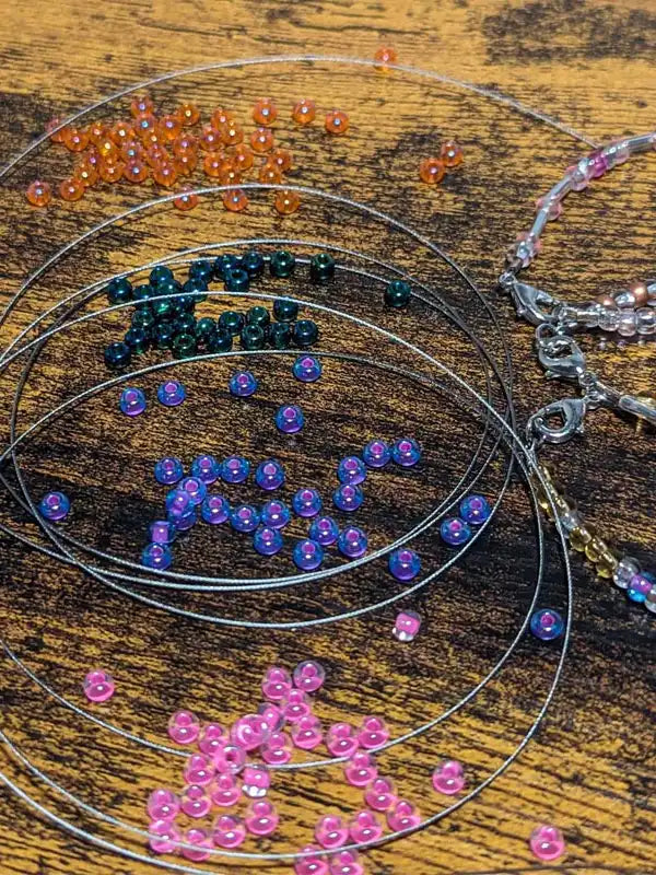A collection of Morse code bracelet crafting materials on a wood tabletop that includes different colored glass beads, stainless steel stringing wire, and completed bracelets.